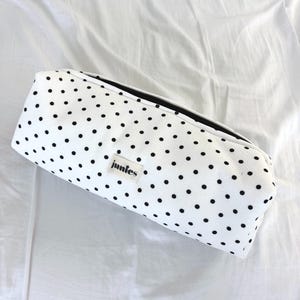 Könnte beinhalten: Weißes Federmäppchen mit schwarzen Punkten. Das rechteckige Etui hat einen schwarzen Reißverschluss und ein kleines Etikett mit der Aufschrift "juntes". Das Etui liegt auf einer weißen Oberfläche.