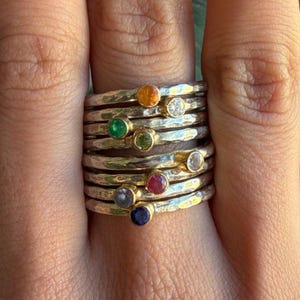 May include: A stack of silver rings with hammered textures, each featuring a different colored gemstone. Gemstones include orange, clear, green, light green, red, light blue, and dark blue. The rings have gold accents around the gemstones.