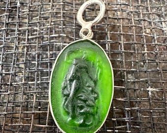 Puede incluir: Un colgante de vidrio verde con una anilla de plata. El colgante presenta una imagen de camafeo de una persona con una túnica fluida.