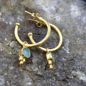 May include: A pair of gold-colored hoop earrings with a small, light blue gemstone pendant. The earrings have a textured finish and a post-and-clasp closure. The gemstone pendants are rectangular with a rough, natural appearance.