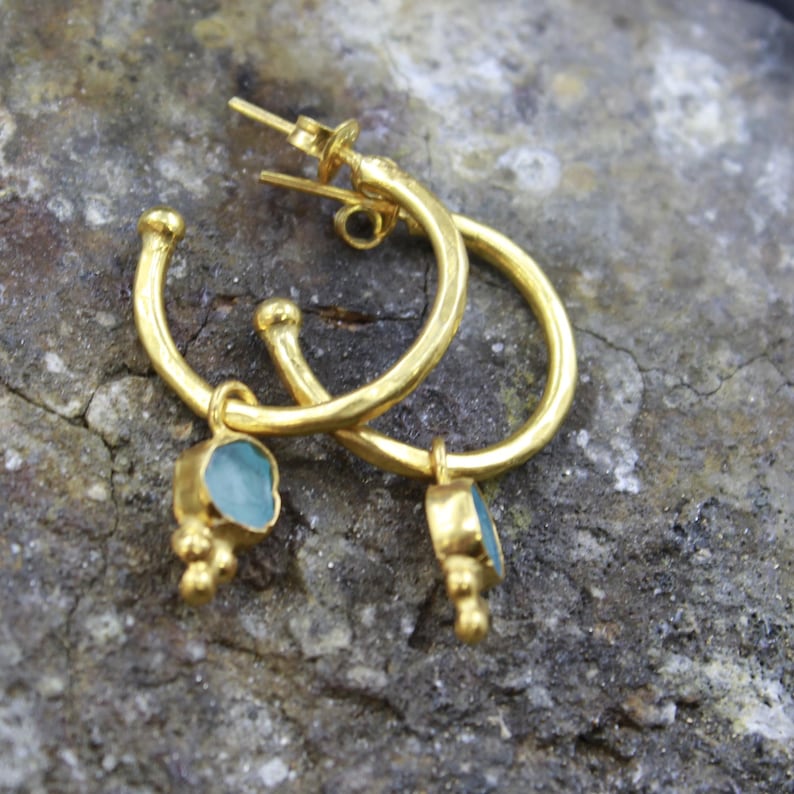 May include: A pair of gold-colored hoop earrings with a small, light blue gemstone pendant. The earrings have a textured finish and a post-and-clasp closure. The gemstone pendants are rectangular with a rough, natural appearance.