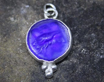 May include: A silver pendant with a round, deep purple glass cameo featuring a raised design of a bird. The pendant has a silver bail and three small silver balls at the bottom.