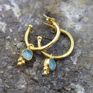 May include: A pair of gold-colored hoop earrings with a small, light blue gemstone pendant. The earrings have a textured finish and are displayed on a rough, gray stone surface. The gemstones have a unique, irregular shape.