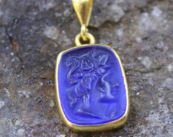 May include: A gold pendant with a rectangular, deep blue stone featuring an engraved classical figure. The pendant has a gold bail for a chain. The stone's design is a detailed relief carving.
