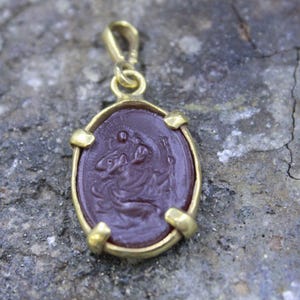 Könnte beinhalten: Ein ovaler Anhänger mit einem tiefroten, durchscheinenden Stein, der in eine goldfarbene Metallfassung mit vier Krappen gefasst ist. Der Stein zeigt ein eingraviertes Relief einer Figur mit Stab und hat eine Öse an der Oberseite.