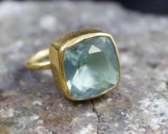 May include: A gold ring featuring a large, square-cut, light blue gemstone. The ring has a simple, classic design with a thick gold band and a bezel setting that holds the gemstone securely. The gemstone has multiple facets.