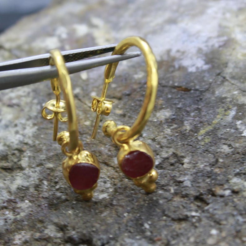Puede incluir: Un par de pendientes de aro dorados con una peque&ntilde;a gema roja y redonda en la parte inferior de cada pendiente. Los pendientes est&aacute;n sujetos por unas pinzas plateadas sobre un fondo gris texturizado.