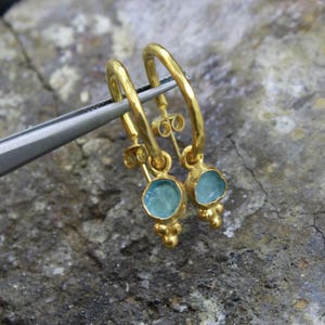 May include: A pair of gold hoop earrings with a small, round, light blue gemstone pendant. The earrings are held by a silver tool against a gray, textured surface. The earrings have a simple, elegant design.