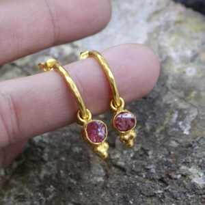 Puede incluir: Un par de pendientes de aro dorados, cada uno con una piedra preciosa redonda de color rosa rojizo. Los pendientes tienen un acabado texturizado y un peque&ntilde;o elemento decorativo debajo.