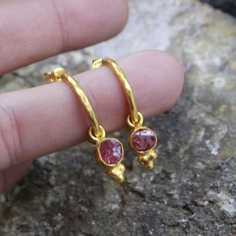 Puede incluir: Un par de pendientes de aro dorados, cada uno con una piedra preciosa redonda de color rosa rojizo. Los pendientes tienen un acabado texturizado y un peque&ntilde;o elemento decorativo debajo.