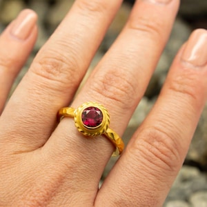 Può includere: Un anello in oro con una pietra preziosa rotonda di colore rosso intenso, incastonata in una cornice a corda intrecciata. Il gambo dell'anello presenta una finitura martellata e testurizzata, in contrasto con la superficie liscia della pietra.