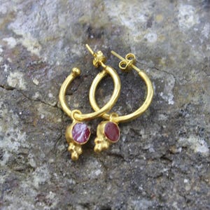 Puede incluir: Un par de pendientes de aro dorados con un peque&ntilde;o colgante redondo de piedra roja. Los pendientes est&aacute;n sobre una superficie gris texturizada. Dise&ntilde;o sencillo y elegante.