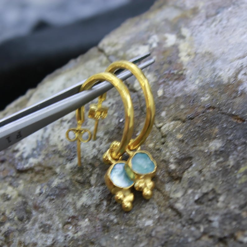 May include: A pair of gold hoop earrings with light blue gemstone pendants. The earrings are held by a silver tweezer against a textured stone surface. The earrings have a unique, handcrafted appearance.