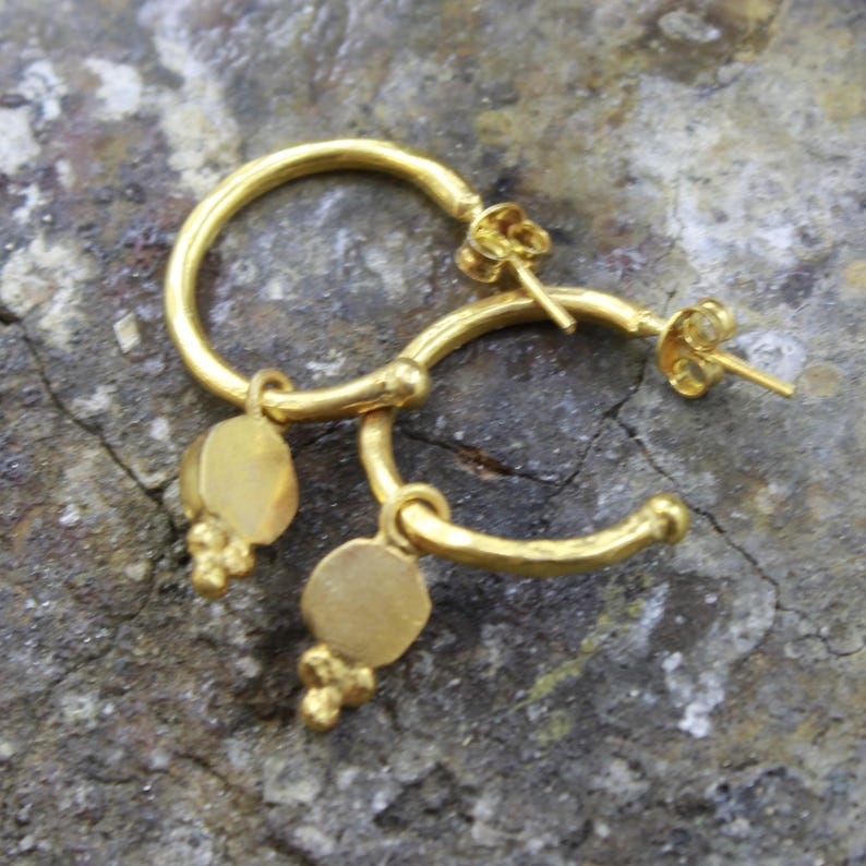 May include: A pair of gold-colored hoop earrings. Each earring has a circular hoop with a small, round charm and a cluster of tiny beads hanging from the bottom. The earrings are displayed on a textured, gray surface.