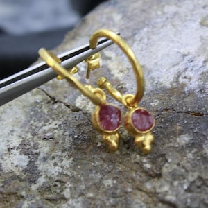 Puede incluir: Un par de pendientes de aro dorados con un peque&ntilde;o colgante redondo de piedra roja. Los pendientes se muestran sobre una superficie gris rugosa, con unas pinzas plateadas.