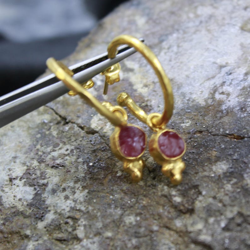 Puede incluir: Un par de pendientes de aro dorados con un peque&ntilde;o colgante redondo de piedra roja. Los pendientes se muestran sobre una superficie gris rugosa, con unas pinzas plateadas.