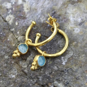 May include: A pair of gold-colored hoop earrings with a light blue gemstone and small gold accents. The earrings are displayed on a textured, gray surface. The earrings have a simple, elegant design.