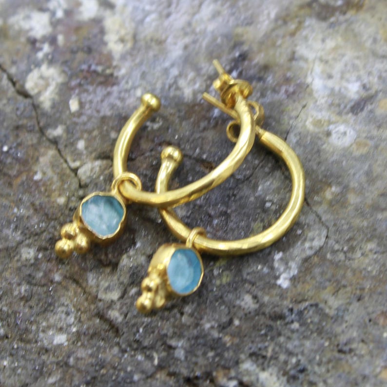 May include: A pair of gold-colored hoop earrings with a light blue gemstone and small gold accents. The earrings are displayed on a textured, gray surface. The earrings have a simple, elegant design.