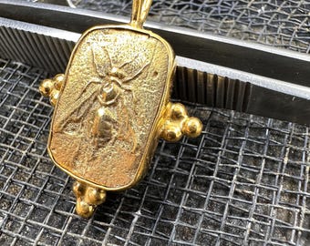 May include: A gold-toned pendant featuring a detailed bee design. The pendant is rectangular with a textured surface and has a loop at the top for attaching to a chain.