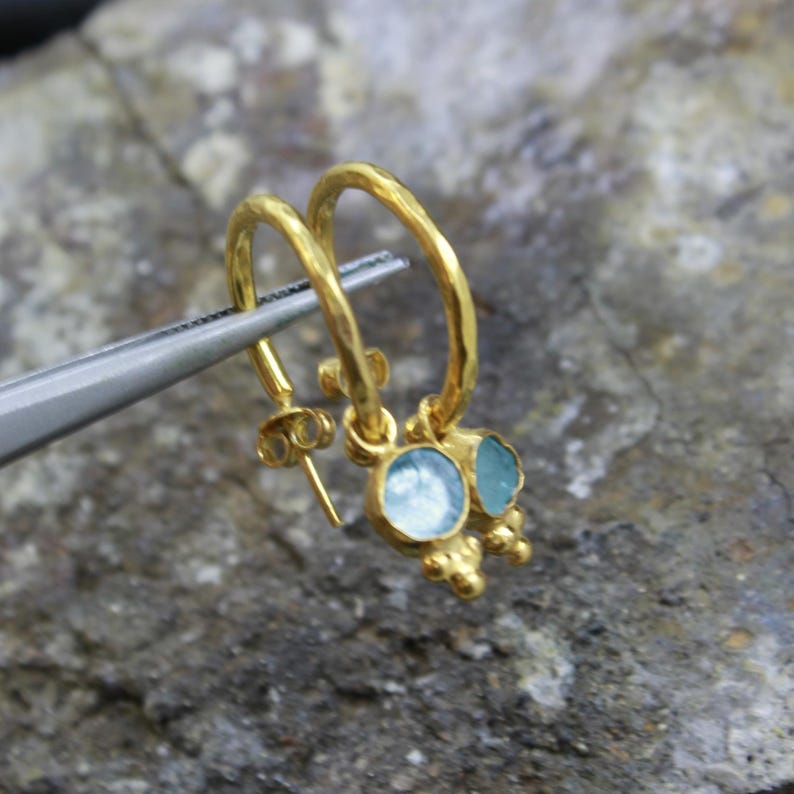 May include: A pair of gold-colored hoop earrings with a hammered texture. Each earring features a light blue gemstone and small, decorative gold beads. The earrings are held by a silver tool against a textured gray background.