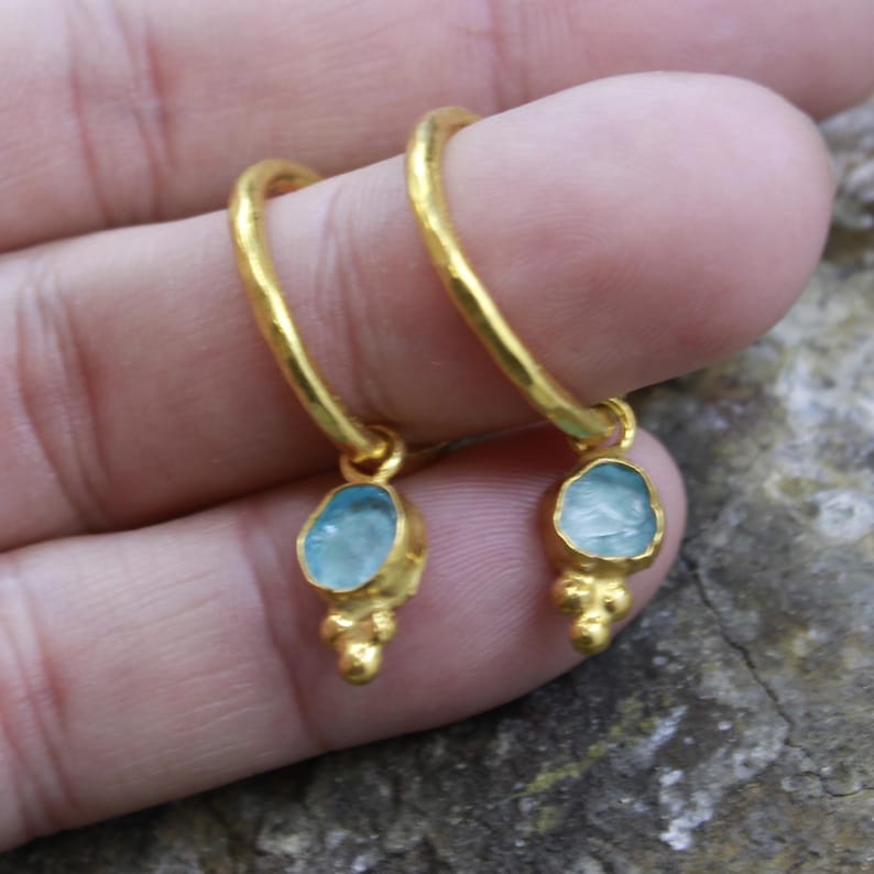 May include: A pair of gold-colored hoop earrings with a light blue gemstone pendant. The pendant has a unique, irregular shape and is attached to the hoop with a small, decorative gold element. The earrings are displayed against a neutral background.