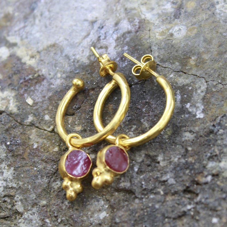 Puede incluir: Un par de pendientes de aro dorados con un peque&ntilde;o colgante redondo de piedra roja. Los pendientes tienen un acabado texturizado y un cierre de poste y tuerca. Las piedras preciosas son de color rojo intenso.