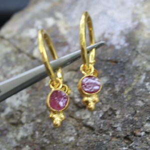 Puede incluir: Pendientes de aro dorados con un peque&ntilde;o colgante redondo de piedra roja. Los pendientes est&aacute;n sujetos por una herramienta plateada sobre un fondo gris texturizado. Los pendientes tienen una textura martillada y un adorno de tres cuentas.