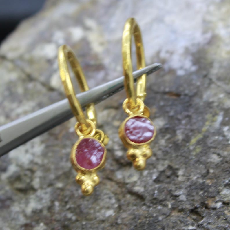 Puede incluir: Pendientes de aro dorados con un peque&ntilde;o colgante redondo de piedra roja. Los pendientes est&aacute;n sujetos por una herramienta plateada sobre un fondo gris texturizado. Los pendientes tienen una textura martillada y un adorno de tres cuentas.