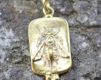 May include: A gold-colored pendant with a rectangular shape and a detailed insect design. The pendant features a loop at the top for a chain and a decorative bottom. The insect is centered on the pendant.