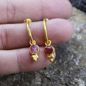 Puede incluir: Un par de pendientes de aro dorados. Cada pendiente presenta una peque&ntilde;a gema rosa de forma ovalada engastada en un bisel dorado. Debajo de la gema hay un grupo de tres peque&ntilde;as cuentas doradas. Los pendientes est&aacute;n sostenidos por una mano.