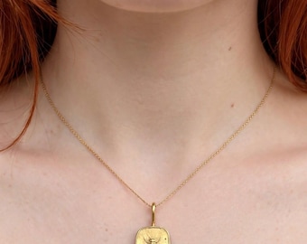 May include: A gold-toned necklace featuring a rectangular pendant with a textured surface and a central embossed bee motif. The pendant has small decorative spheres on the sides and bottom, suspended from a delicate gold-toned chain link necklace.