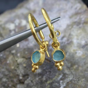 May include: A pair of gold-colored hoop earrings with a light blue stone pendant. Each earring has a small cluster of gold beads below the stone. The earrings are held by a silver tool.
