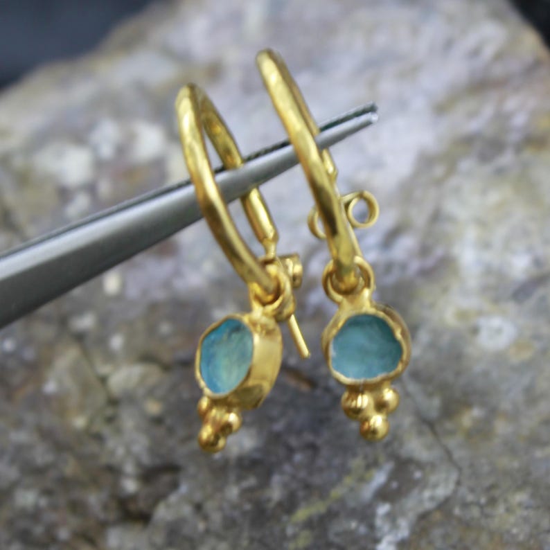 May include: A pair of gold-colored hoop earrings with a light blue stone pendant. Each earring has a small cluster of gold beads below the stone. The earrings are held by a silver tool.
