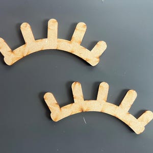 Op de afbeelding: Twee houten uitsnijdingen van wimpers, elk met een gebogen vorm en vijf punten aan de bovenkant. De wimpers zijn lichtbruin.