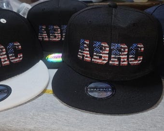 Op de afbeelding: Vier zwarte baseballpetten met witte kleppen. Elke pet heeft een ander Amerikaans vlagontwerp met de letters "ABRC" op de voorkant. De petten hebben een "Snapback"-label op de achterkant.