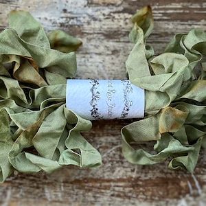 May include: Two bundles of olive green fabric ribbon with frayed edges. The ribbon is wrapped around a white label with the text "Land Rose Studios" in a decorative font. The ribbon is on a weathered wooden surface.