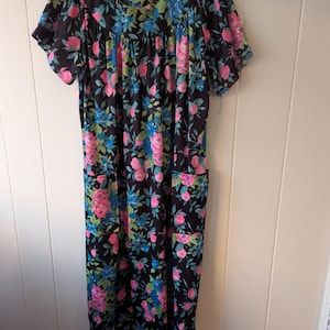 Puede incluir: Vestido floral negro con rosas rosas y azules y hojas verdes. El vestido tiene mangas cortas, cuello redondo y dos bolsillos. Cuelga de una percha blanca.
