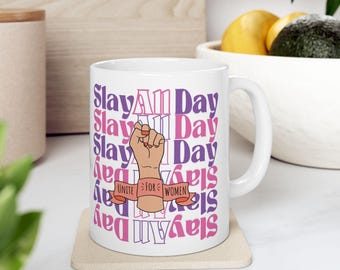 May include: White ceramic mug with a pink and purple graphic that says "Slay All Day" and features a raised fist with a pink wristband that says "Unite For Women".