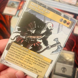 Könnte beinhalten: Eine Fantasy-Sammelkarte mit Kratos, Gott des Krieges, einer legendären Kreatur mit einer rot-schwarzen Axt. Die Karte hat einen gelben Hintergrund mit schwarzem Text und einem weißen Rand. Der Text lautet "Kratos, Gott des Krieges (Kardik, Doon Source)" und "Legendäre Kreatur - Dämonen-Berserker".