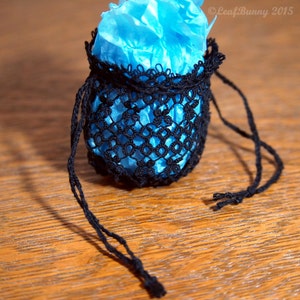 May include: A small black crocheted drawstring bag with a blue tissue paper inside. The bag has a long black cord that can be tied to close the bag.