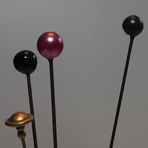 May include: A collection of decorative hat pins with various designs. The pins feature a black, a pink, and a black ball-shaped top, and a gold-colored, saucer-shaped top. The pins have long, slender, dark metal stems.