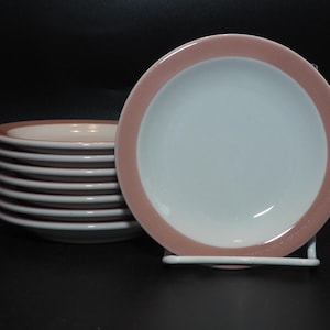 May include: A stack of white plates with a dusty rose border, next to a single plate in a white metal stand. The plates have a simple, clean design, suitable for everyday use. The plates are on a black surface.