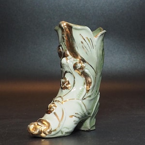 May include: A vintage, light green ceramic boot-shaped vase with gold accents. The boot features floral and leaf designs, with gold trim along the edges and details. The heel and toe are also highlighted in gold.