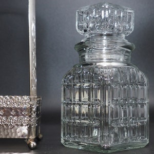 May include: A clear glass bottle with a square, textured design. The bottle has a square lid with a similar textured design.