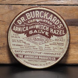 May include: A round, white tin with brown lettering. The tin is labeled "Dr. Burckard's Carbolated Arnica and Witch Hazel Salve". The text also says "For Man and Beast" and "Safe-Effective-Harmless". The tin is made by The American Products Co. in Cincinnati, Ohio, USA.