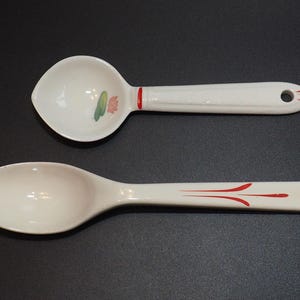 Op de afbeelding: Twee witte keramische lepels. Eén is een serveerlepel met een afgeronde kom en een bloemmotief. De andere is een sleuflepel. Beide hebben rode accentlijnen op de handvatten en een gat om op te hangen.