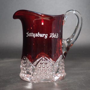 May include: A red glass pitcher with a clear glass handle. The pitcher has a clear glass base with a cut glass design. The pitcher is etched with the words "Gettysburg 1863".