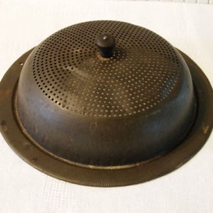May include: A vintage metal colander with a round, perforated lid and a black knob on top. The colander is made of a dark metal and has a slightly worn appearance.