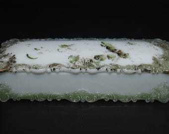 May include: A white porcelain trinket box with a green and gold floral design. The box has a rectangular shape with a raised, ornate lid.