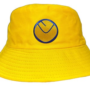 Puede incluir: Un gorro de pescador amarillo con un logotipo bordado en azul y amarillo. El logotipo es un círculo con una letra V en su interior.